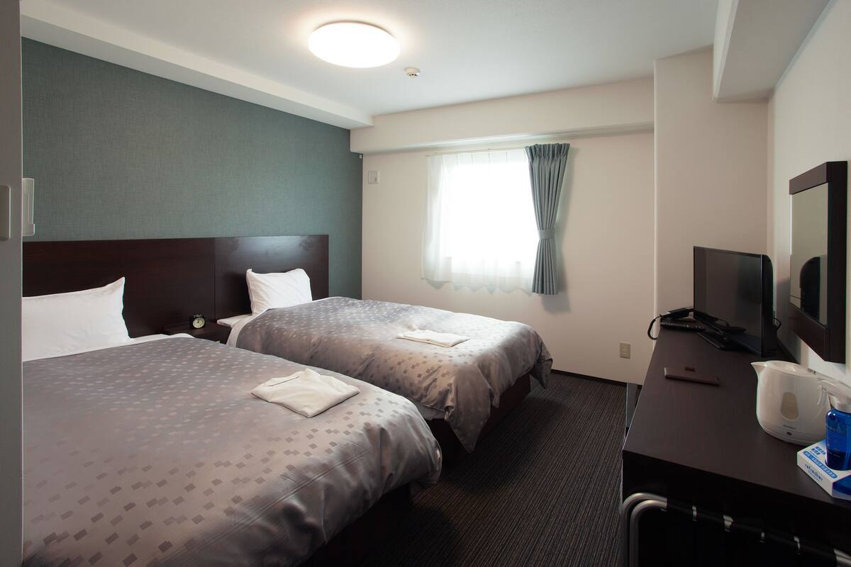 Ein Hotelzimmer mit zwei Betten, einem Schreibtisch mit Fernseher und einem Fenster mit Vorhängen.