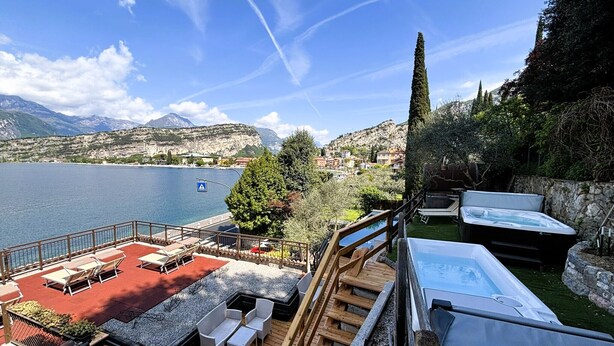 A deck with outdoor furniture, a hot tub, and a view of a lake and mountains. - Hotel Paradiso (Nago-Torbole)