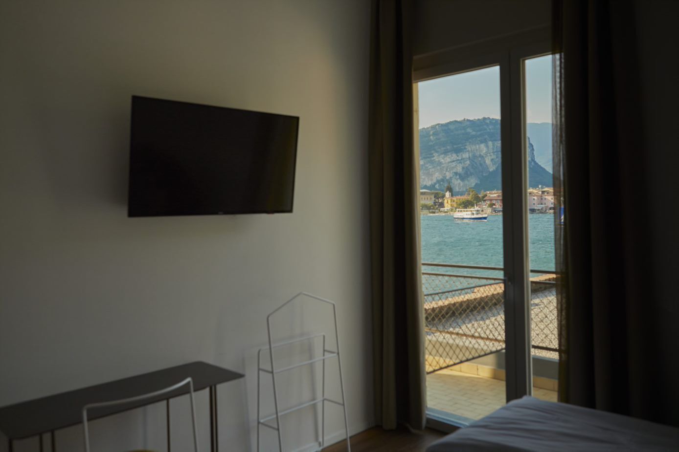 A hotel room with a bed, a TV mounted on the wall, and a view of a body of water with boats and buildings in the distance.