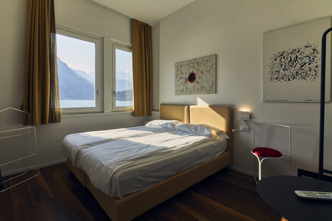 A hotel room with a bed, a window with curtains, a bedside table with a lamp, a chair, and artwork on the wall.