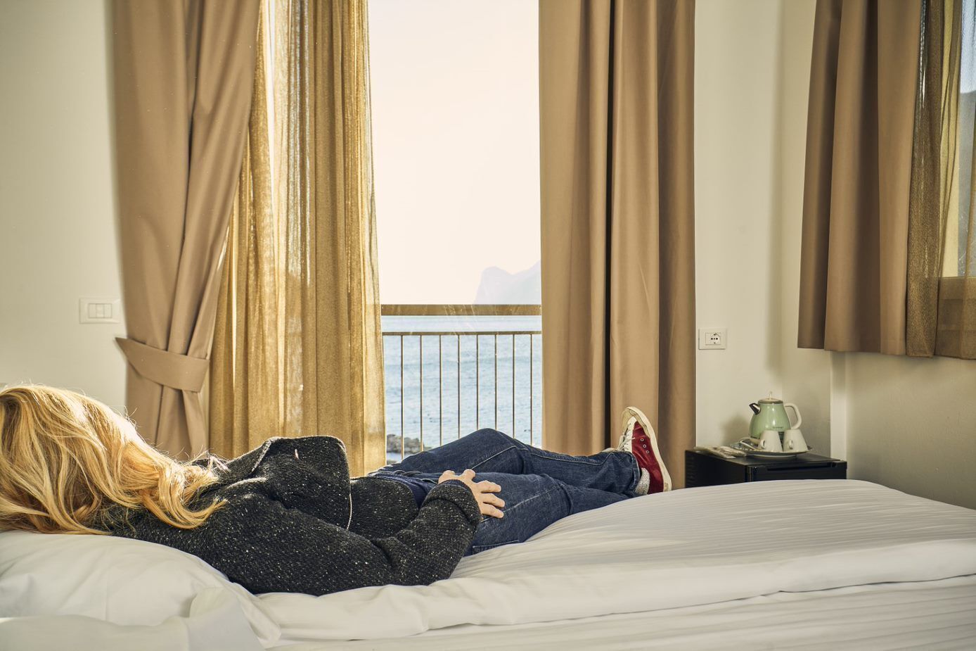A person lying on a bed in a room with a view of the sea through large windows, curtains, and a small table with a teapot.