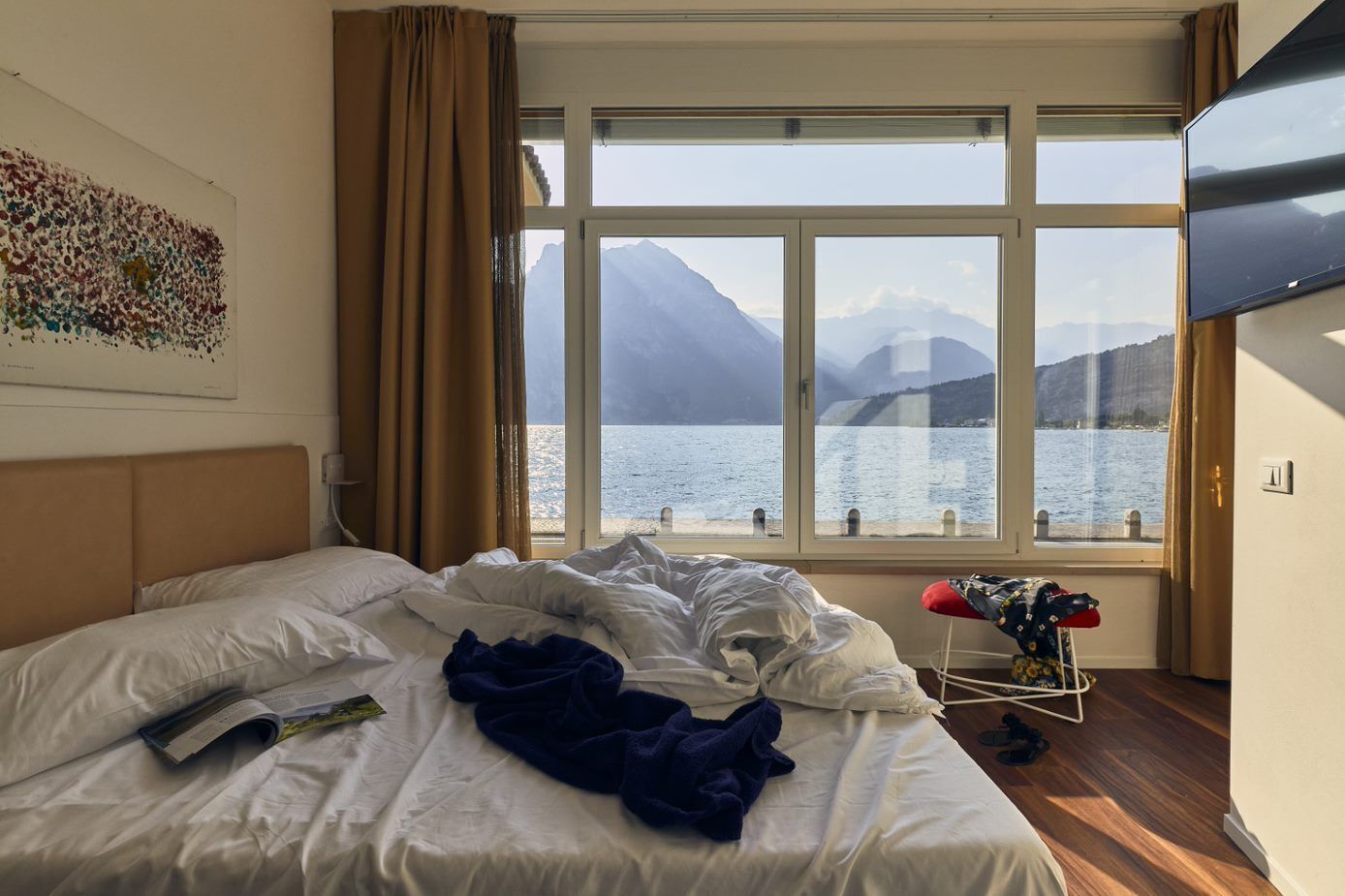 A hotel room with a bed, a window with a view of a lake and mountains, and a TV on the wall.