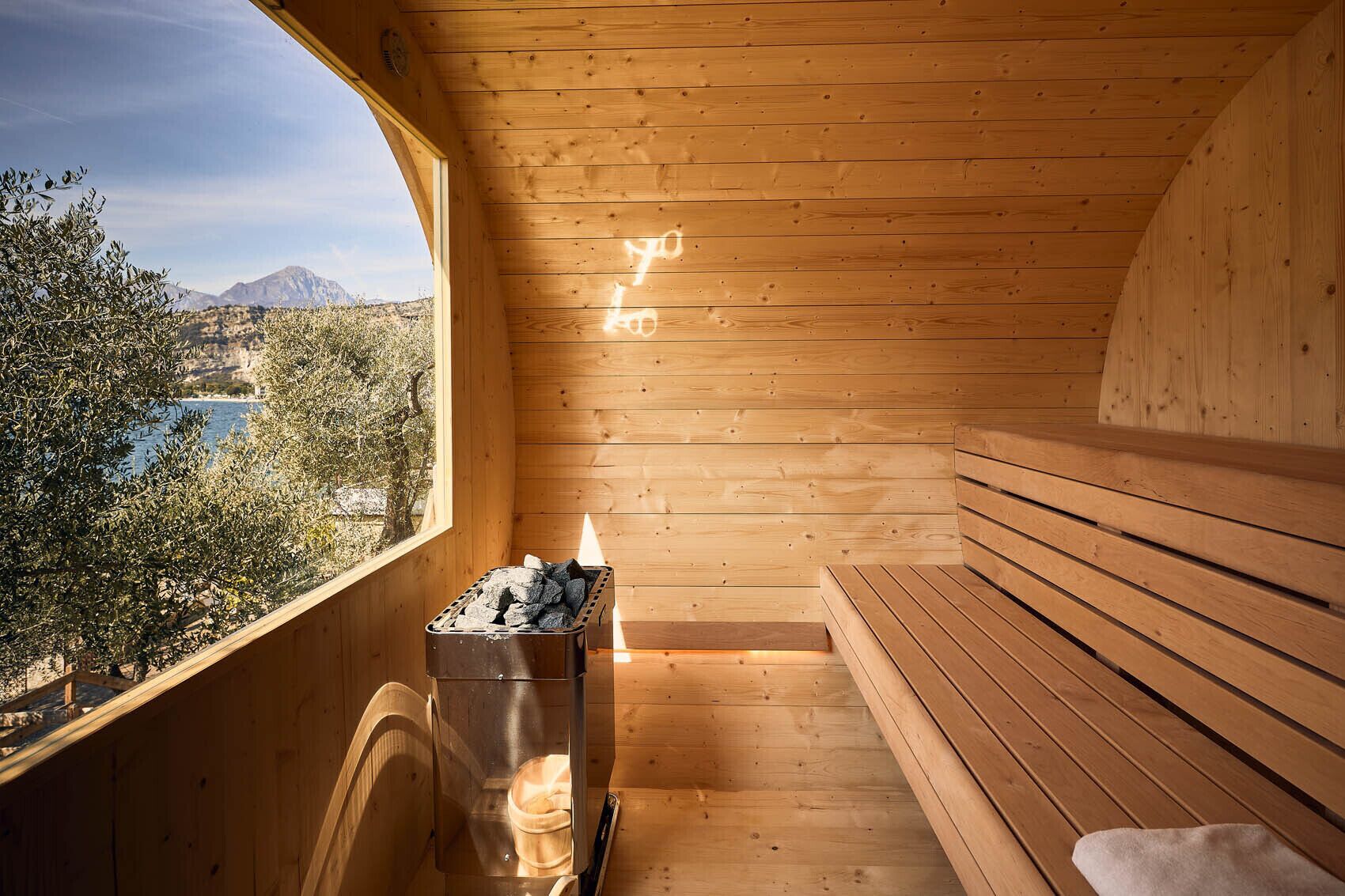 A wooden sauna with a bench, a sauna heater, and a view of a lake and mountains.