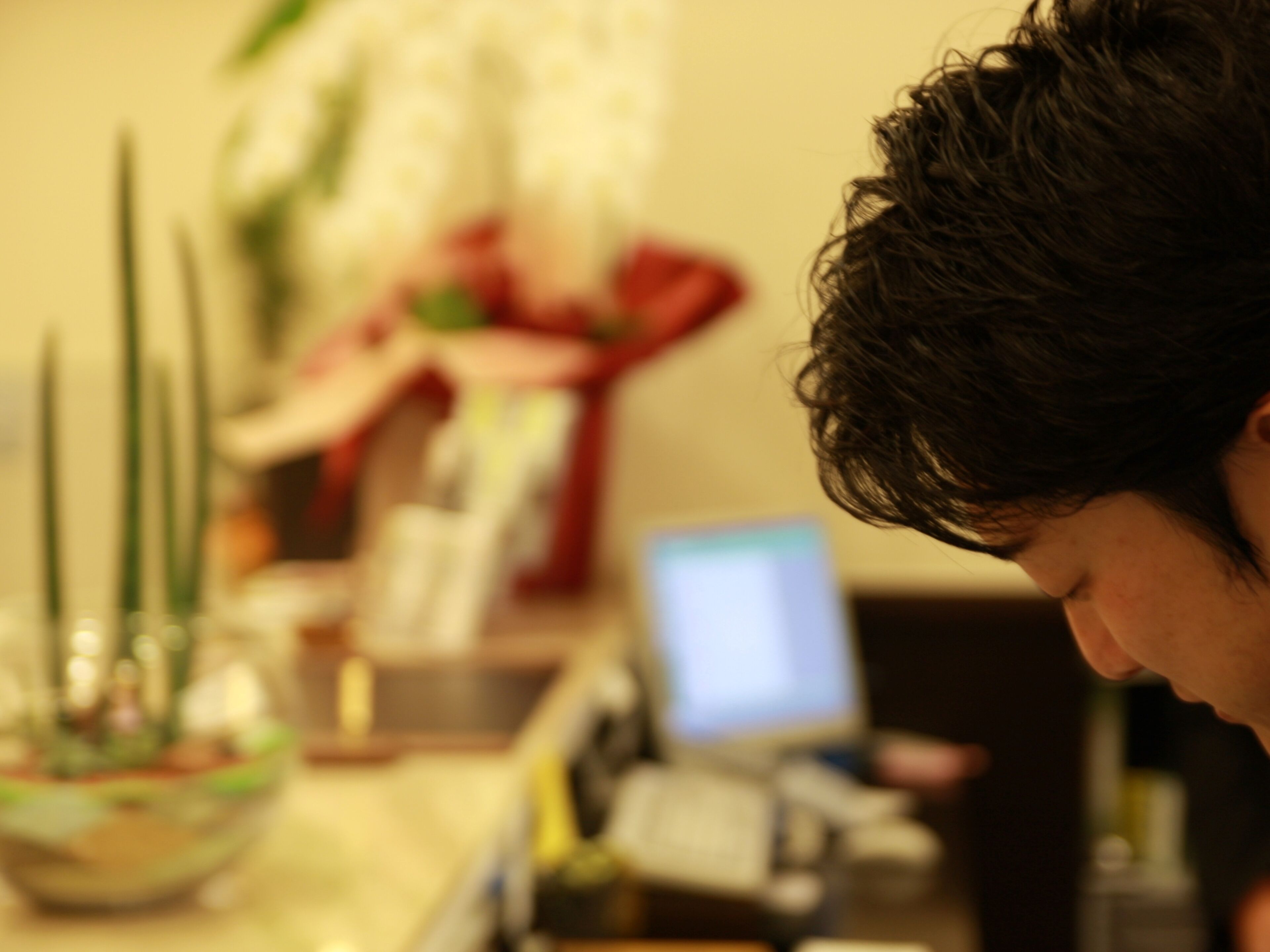 A person sitting at a table with a computer monitor, a potted plant, and a vase with flowers.