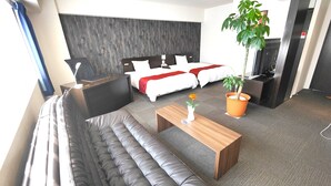 Una habitación de hotel moderna con un sofá grande de color gris, una mesa de centro de madera, una planta en maceta y una cama con ropa de cama blanca y detalles rojos.