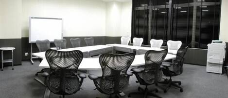 A conference room with a long table, four black mesh chairs, a whiteboard, and a large window with blinds.