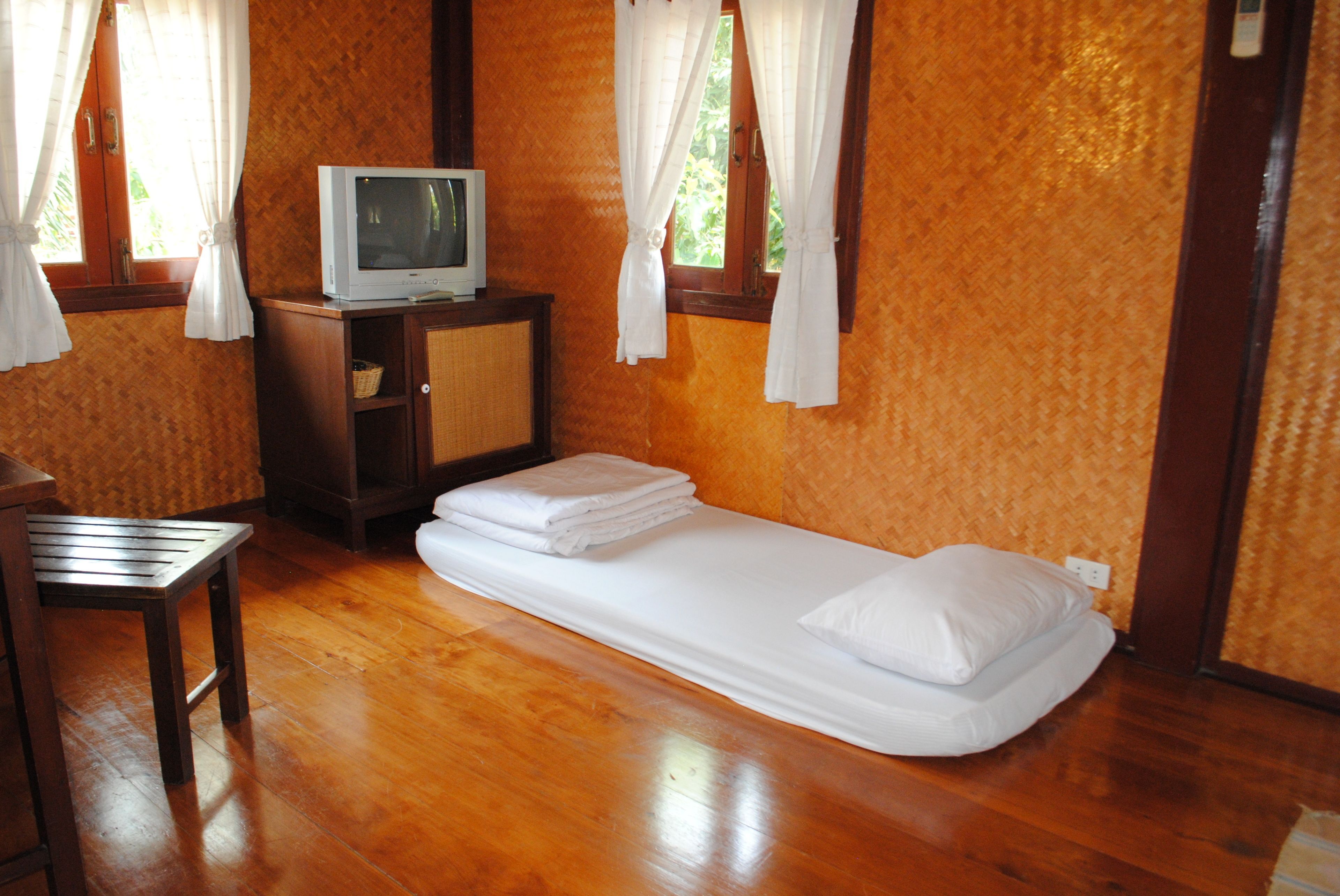 A room with a wooden floor, a single bed with white bedding, a small wooden table, a television on a cabinet, and windows with white curtains.