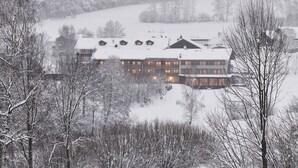 Un edificio grande de varios pisos con tejado cubierto de nieve, rodeado de árboles y un paisaje montañoso.