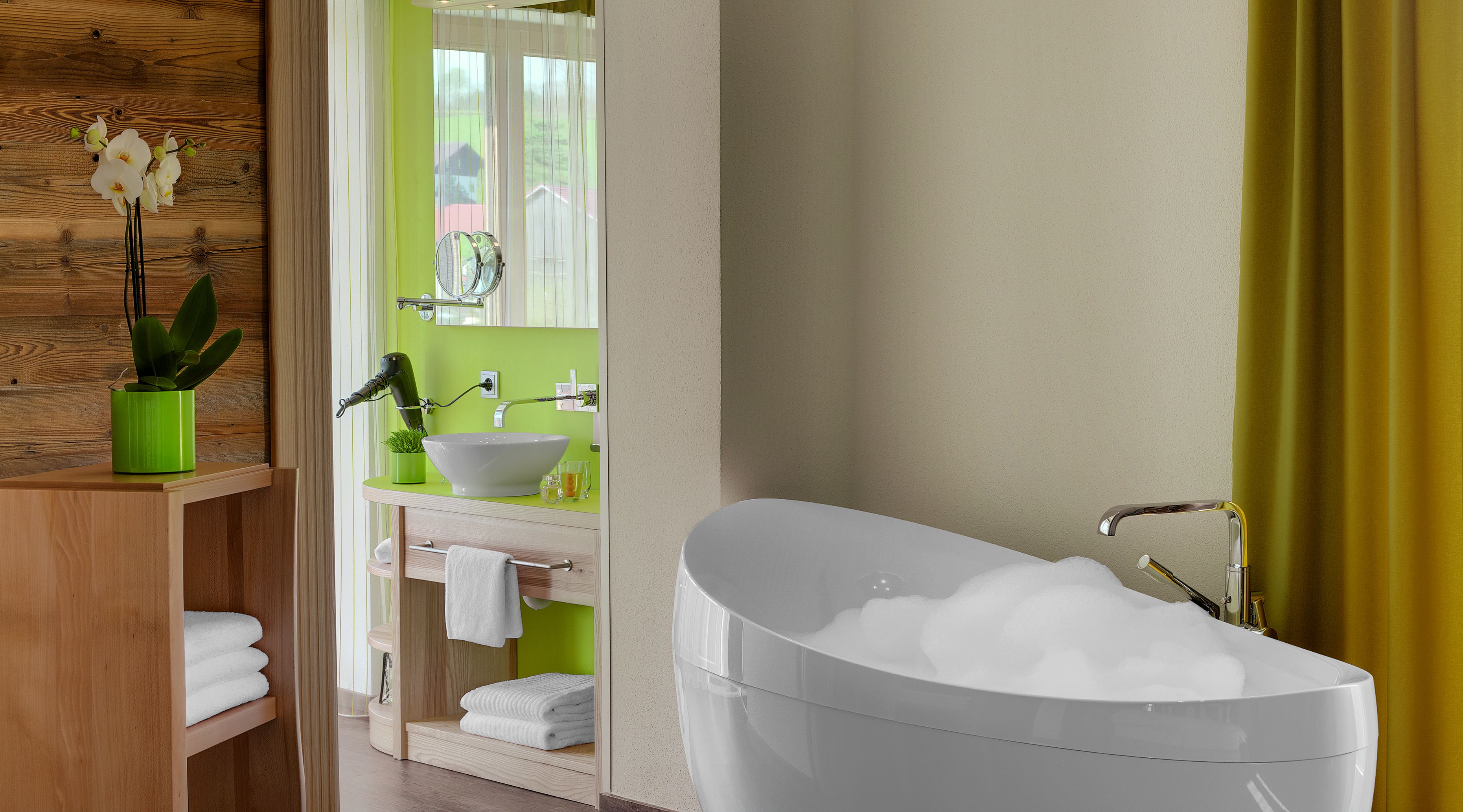 Een moderne badkamer met een vrijstaand bad, een groene wastafelmeubel en een houten plank met een plant.