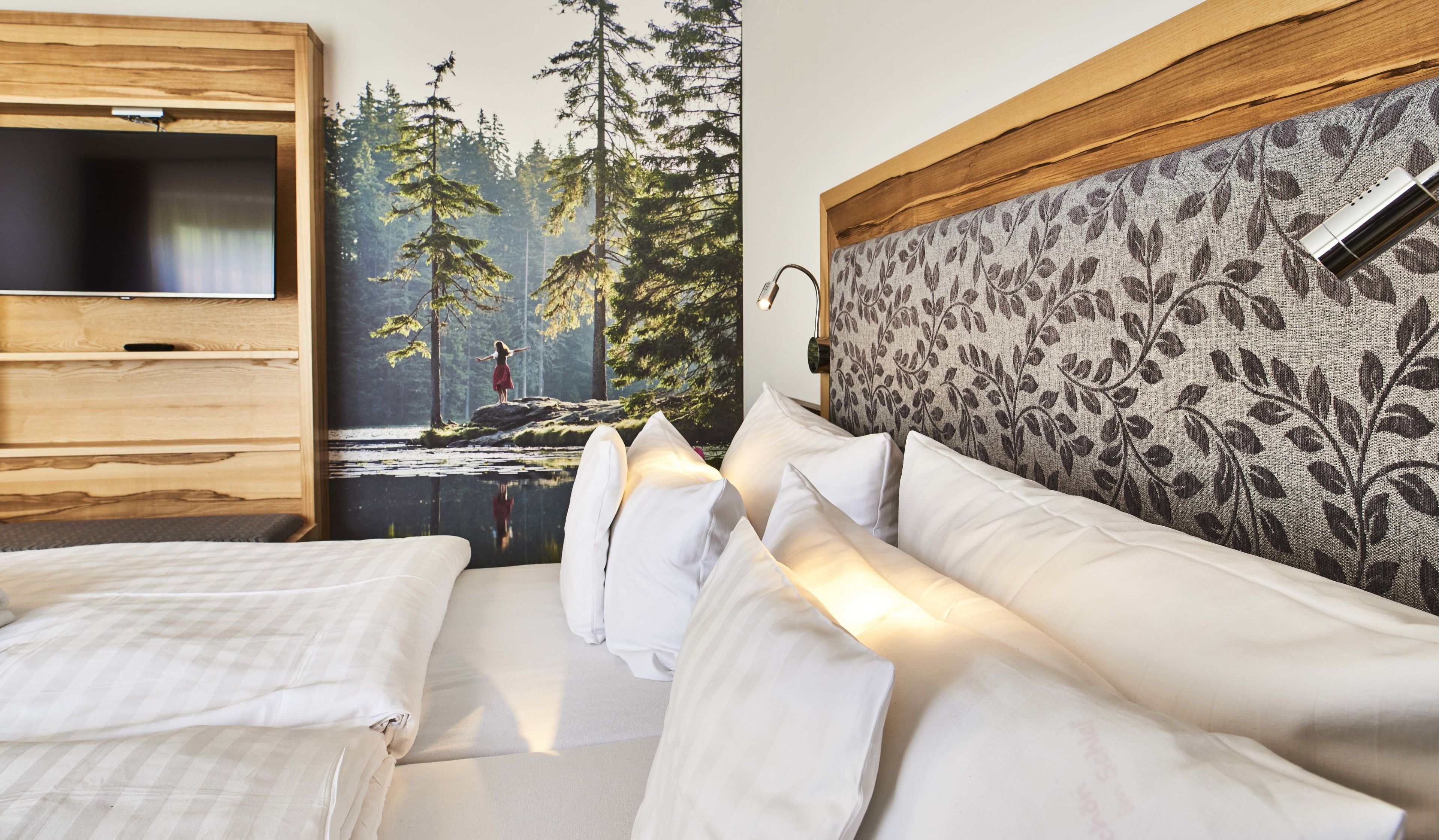 A bedroom with a bed, a TV mounted on a wooden panel, and a forest mural on the wall.