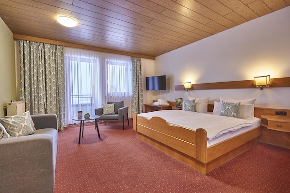 Quarto de hotel com cama de madeira, sofá, mesa pequena e TV fixada na parede.