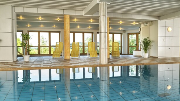 Una amplia piscina interior con azulejos azules cristalinos, sillones de relax amarillos y grandes ventanales con vistas a zonas verdes.