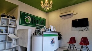 Zona de recepción de un hotel con una pared verde, una araña, un mostrador de recepción, un televisor y dos sillas rojas.