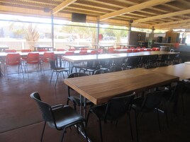 Un área de comedor al aire libre con techo, varias mesas y sillas, una barra larga y un altavoz montado en el techo.