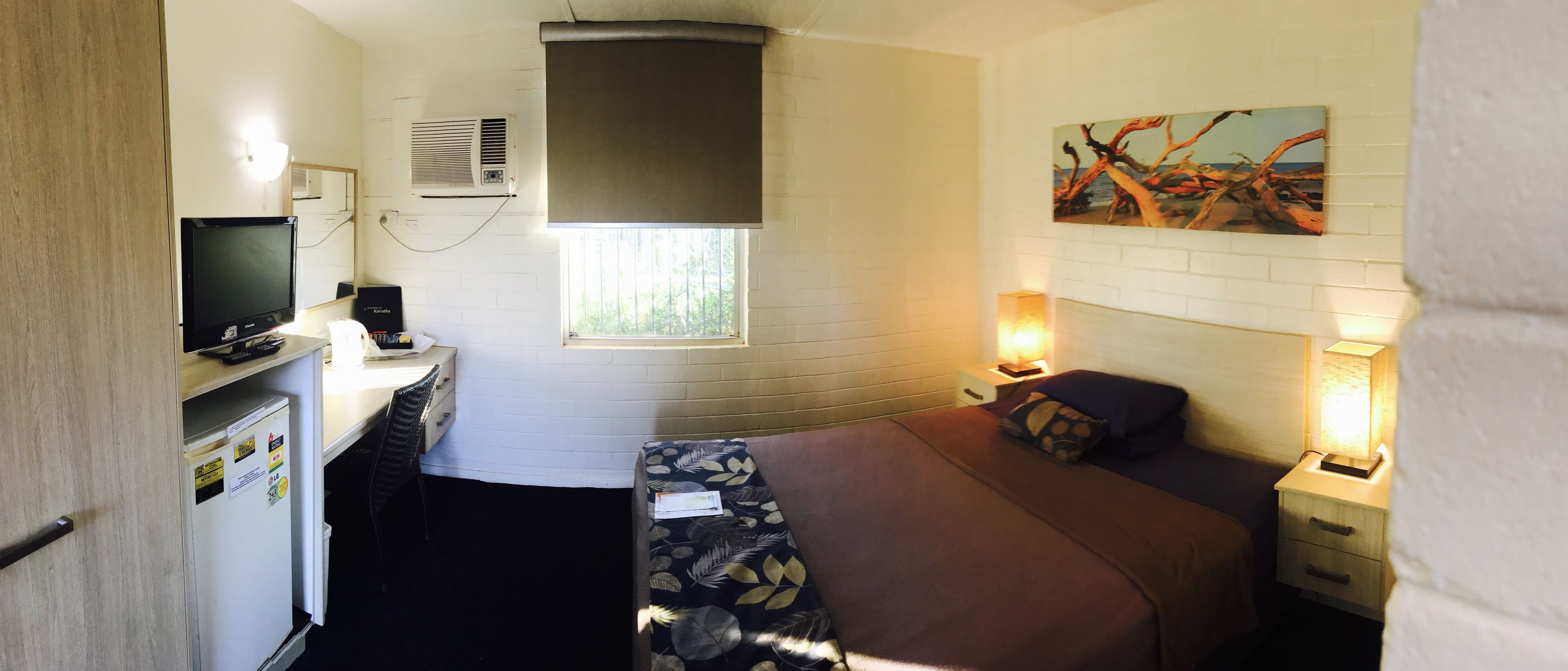 Habitación de hotel pequeña con cama, mesita de noche, lámpara, escritorio con computadora y un refrigerador compacto.