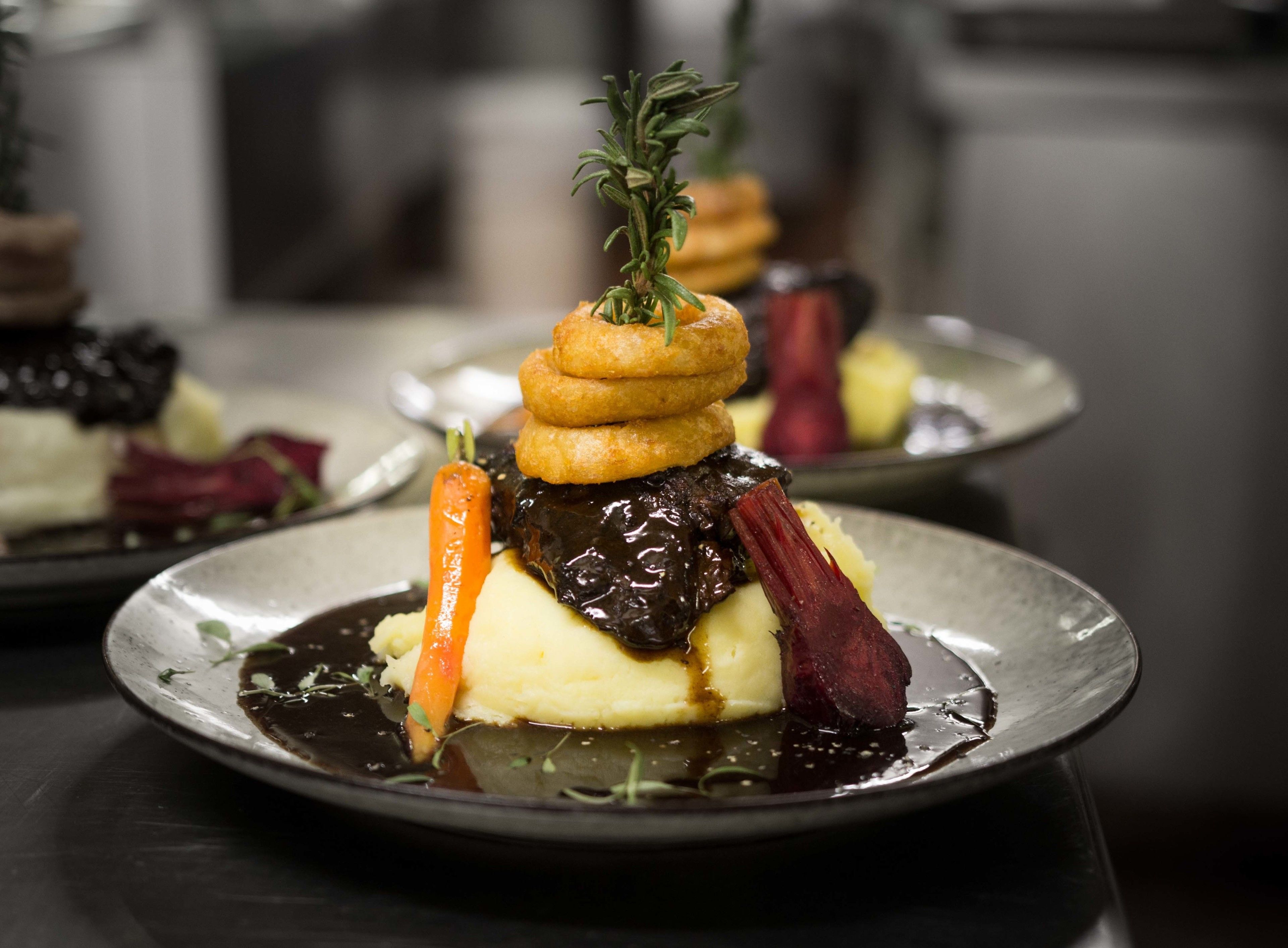Een bord met gepureerde aardappelen, een wortel en een rood groentestuk, mogelijk biet, overgoten met een donkere saus en versierd met een takje rozemarijn.