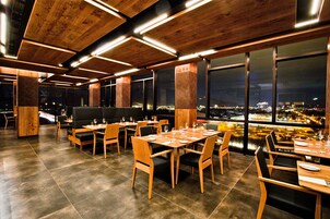 Un restaurante moderno con mesas y sillas de madera, ventanas amplias con vista a la ciudad y un techo alto con vigas a la vista.