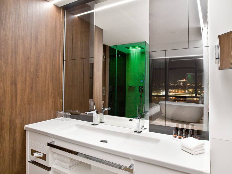 A modern bathroom with a large mirror, a white vanity, and a view of a cityscape through a window.