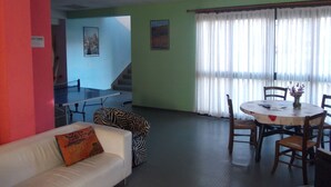 Een kamer met een witte bank, een stoel met zebraprint, een eettafel met stoelen, een trap en een groot raam met dunne gordijnen.