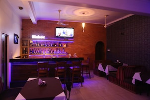 A bar area with a brick wall, a television mounted above the bar, and a seating area with tables and chairs.