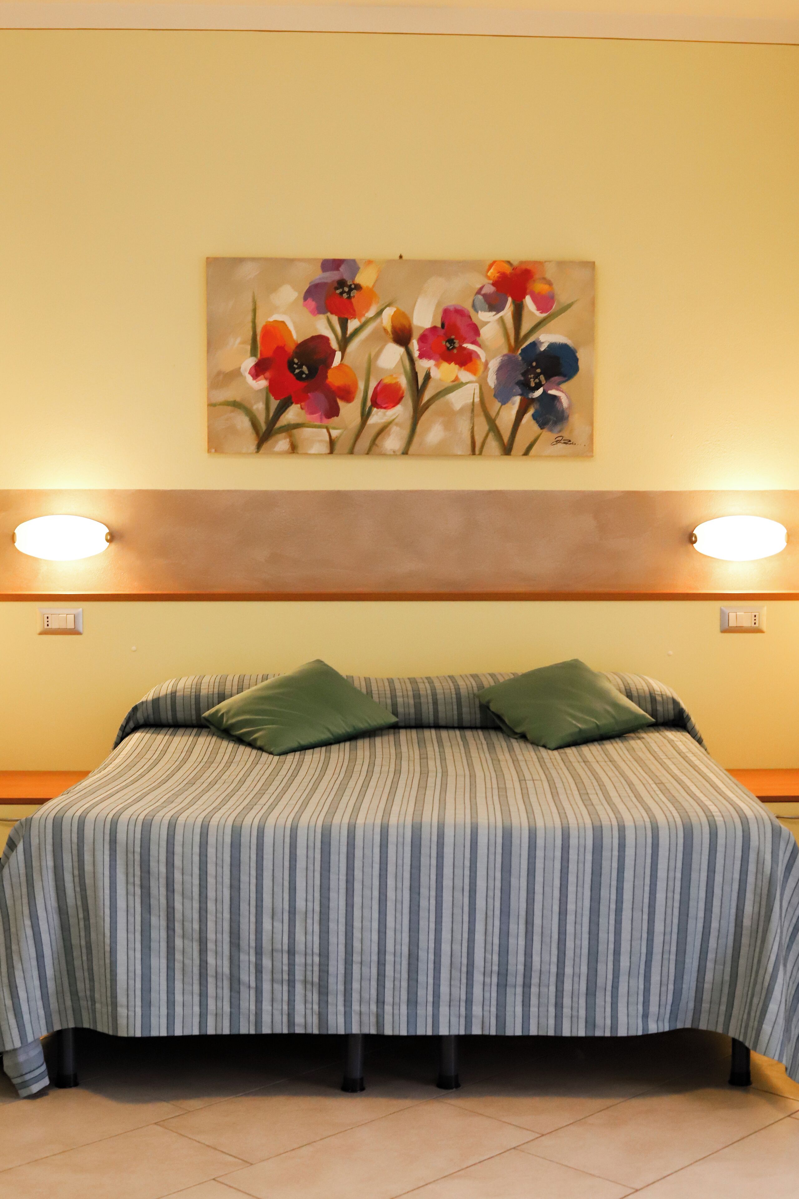 Een hotelkamer met een bed, gestreept beddengoed, twee groene kussens, een bloemen schilderij aan de muur en wandlampen.