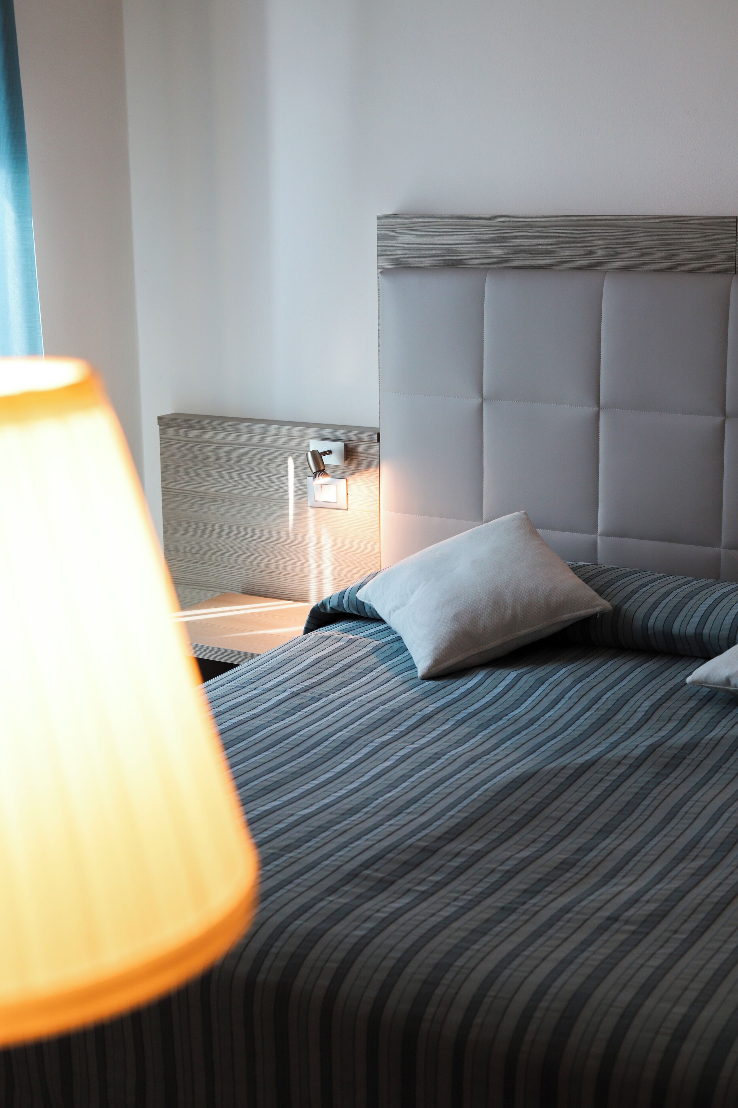 A neatly made bed with striped bedding, a headboard with a light wood finish, and a bedside lamp.