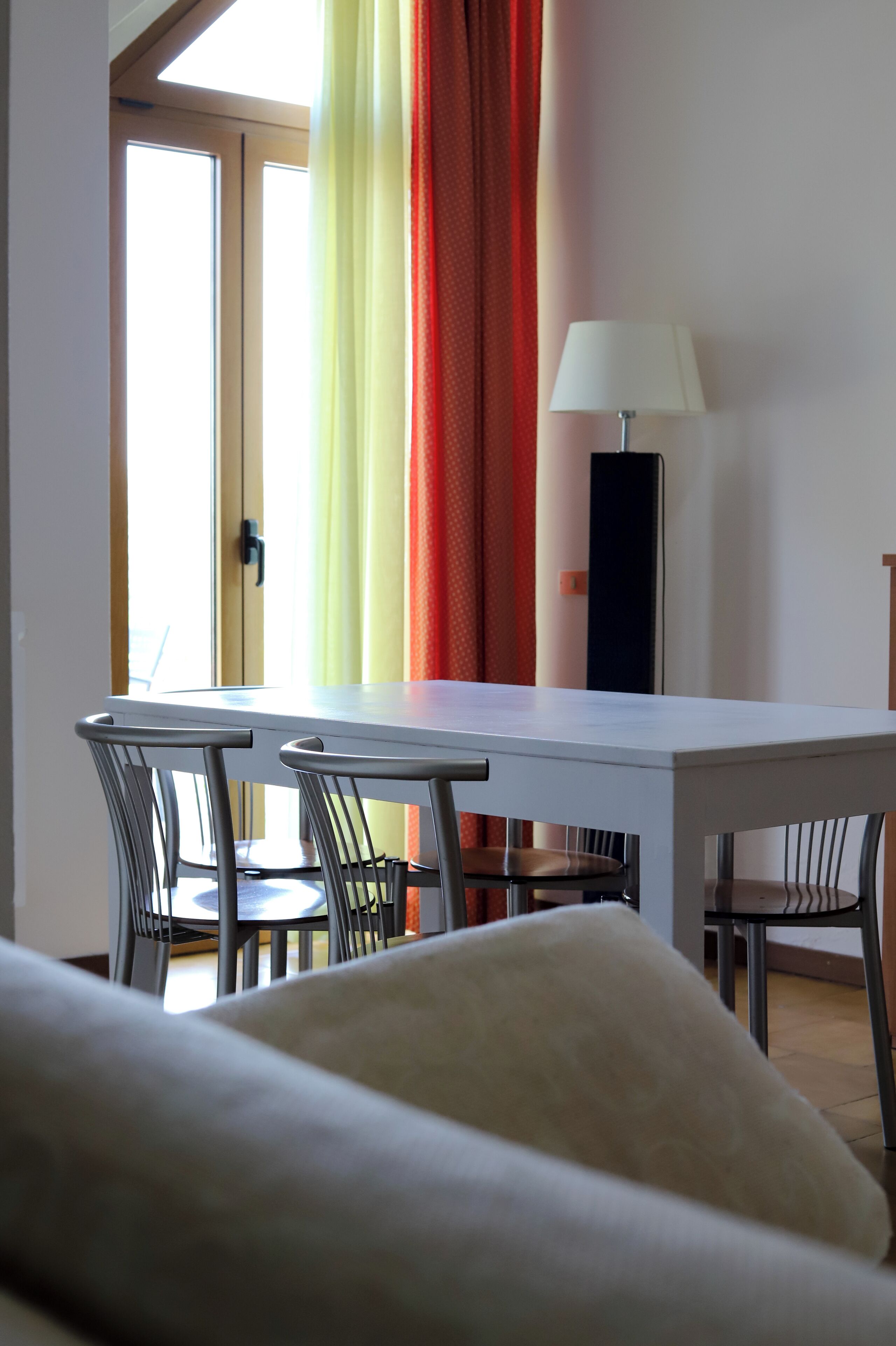 Uma área de refeições com mesa branca e quatro cadeiras, luminária e vista pela janela com cortinas.