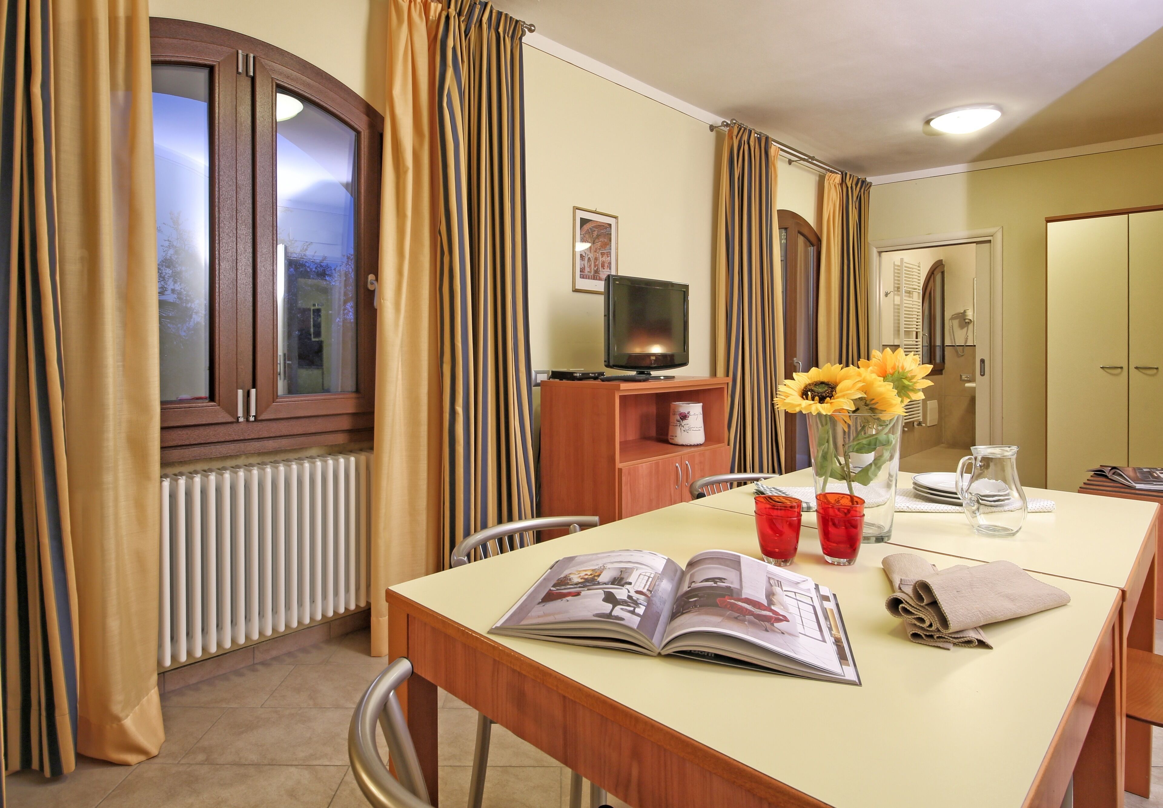 A dining area with a table, chairs, a television, a radiator, curtains, a vase with flowers, and a magazine.