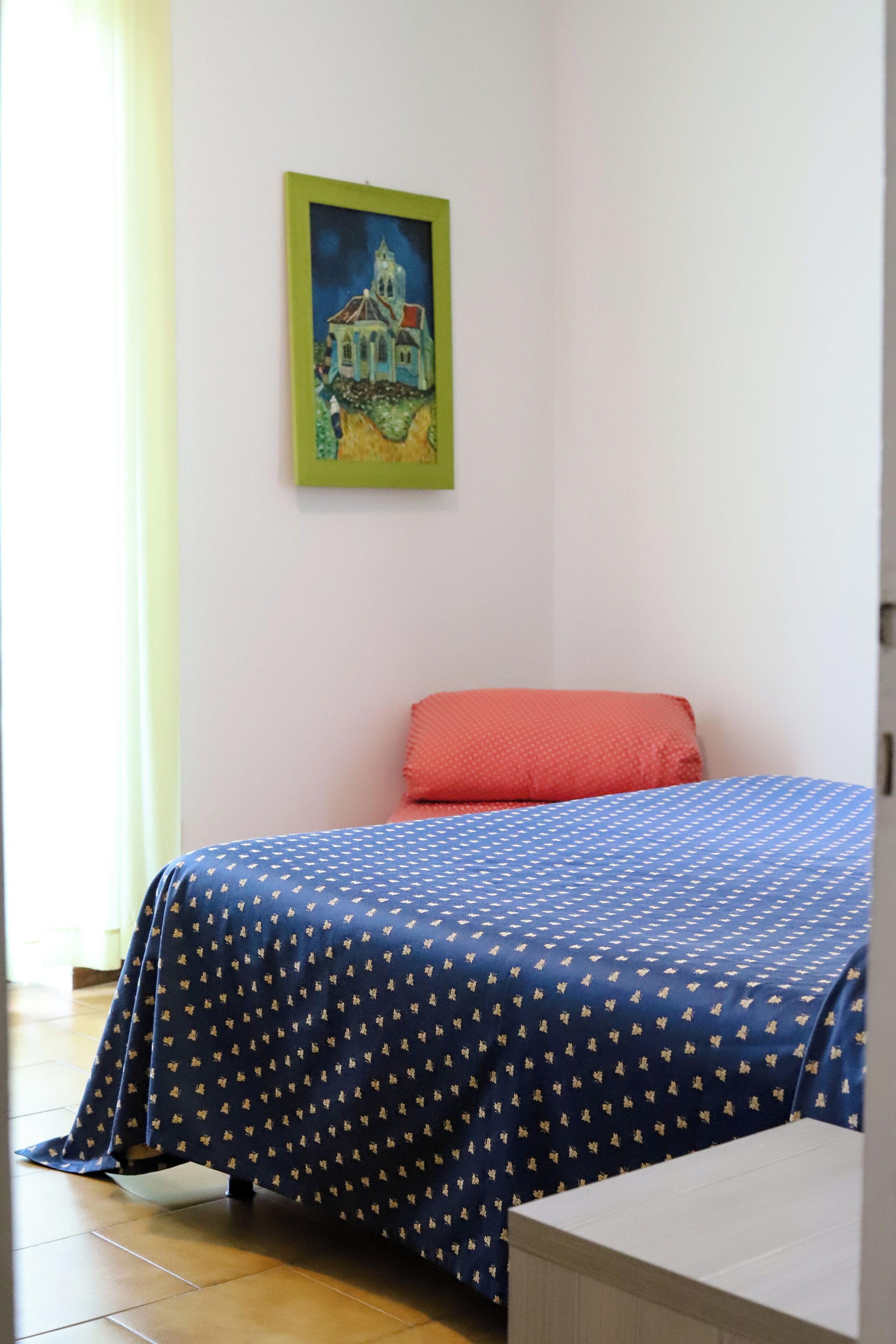 Un dormitorio con una cama, un almohadón rojo, una colcha azul con estampado y un cuadro en la pared.