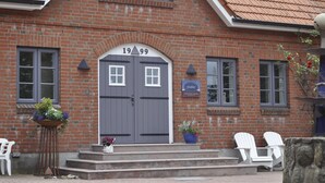 A brick building with a grey double door, two windows, and a small outdoor seating area with white chairs. - urlaubsart - Guldehof (Stoltebuell)