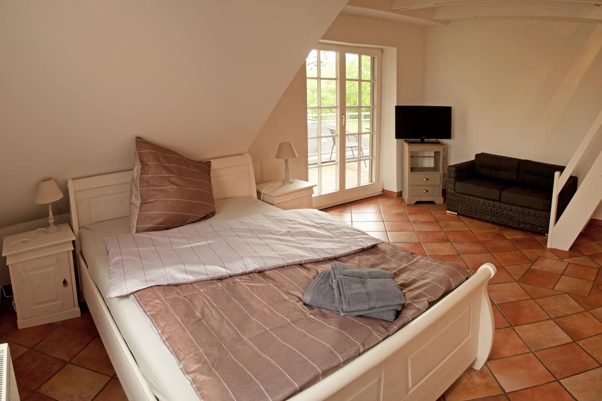 A bedroom with a bed, a nightstand, a TV, a sofa, and a window.