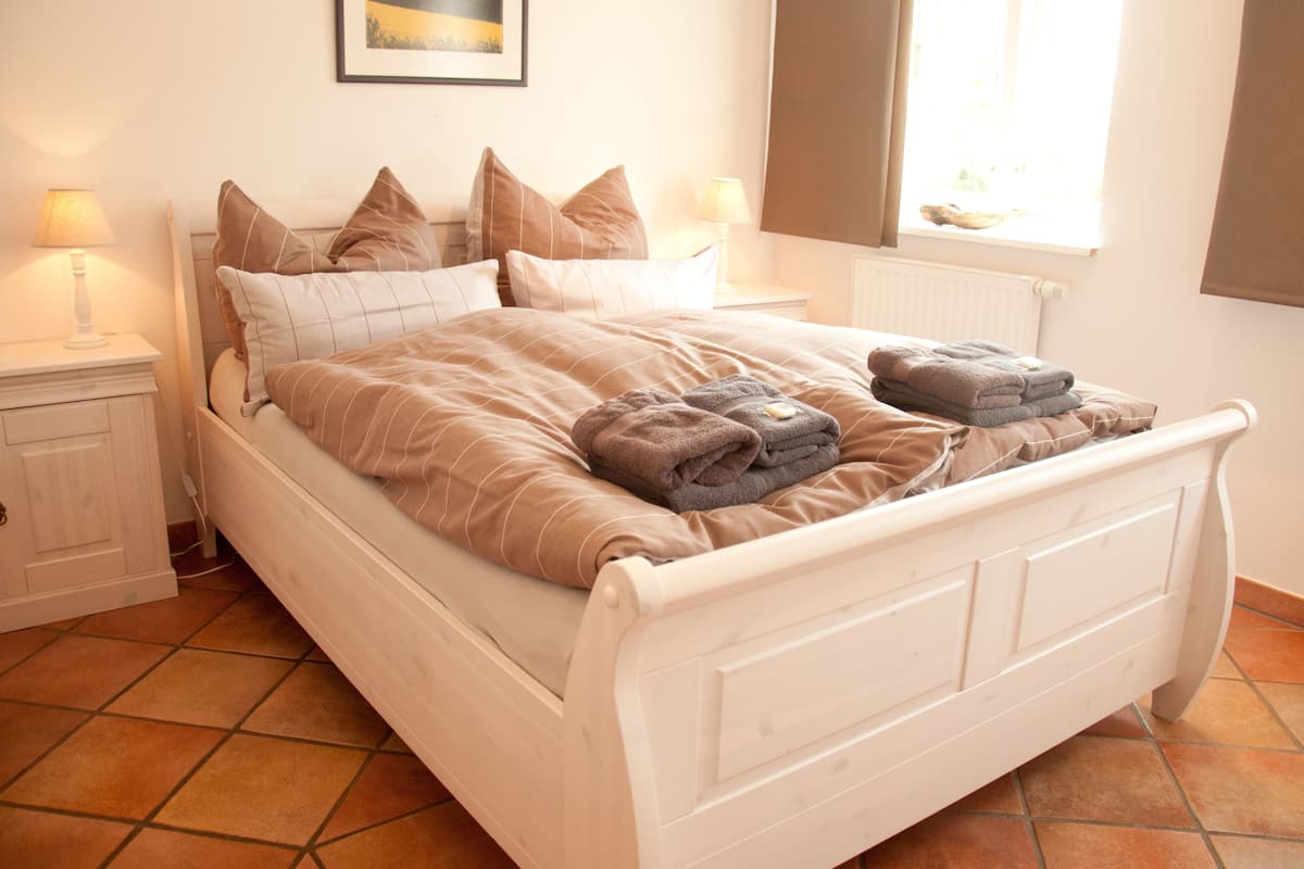 A neatly made bed with a brown comforter and pillows, a bedside table with a lamp, and a window with curtains.