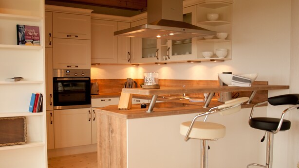 A modern kitchen with white cabinets, a built-in oven, a stainless steel range hood, and two bar stools. - urlaubsart - Guldehof (Stoltebuell)