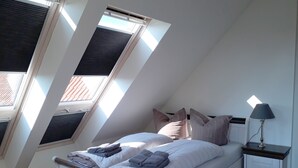 A bedroom with a sloped ceiling, skylights, a bed with pillows, a bedside table with a lamp, and a small cabinet. - urlaubsart - Am Rathaus (Kappeln)