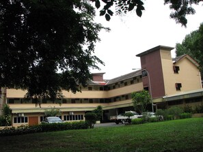 A multi-story building with a flat roof, surrounded by greenery and parked vehicles.
