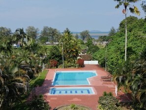 A swimming pool with a red border, surrounded by palm trees and greenery, with a view of the sea and mountains in the background.
