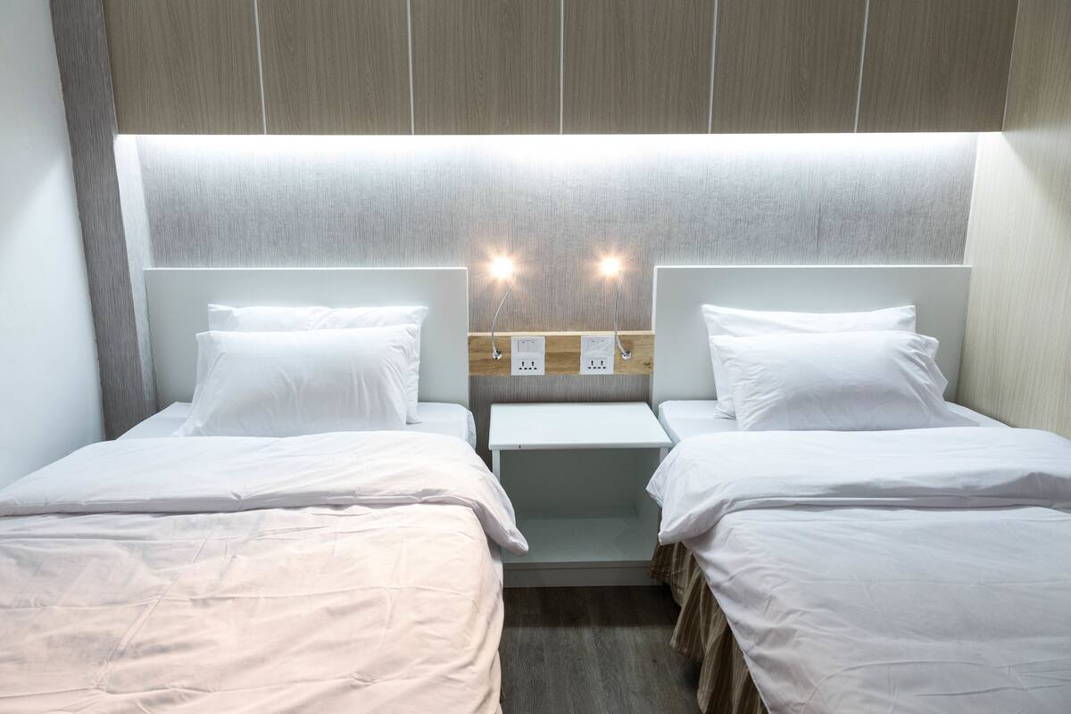 A hotel room with two single beds, a small bedside table, and a wall-mounted light fixture.