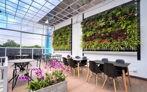 A modern indoor space with a glass ceiling, a variety of potted plants on a vertical garden wall, and a seating area with tables and chairs. - Connect Hostel (Chiang Rai)
