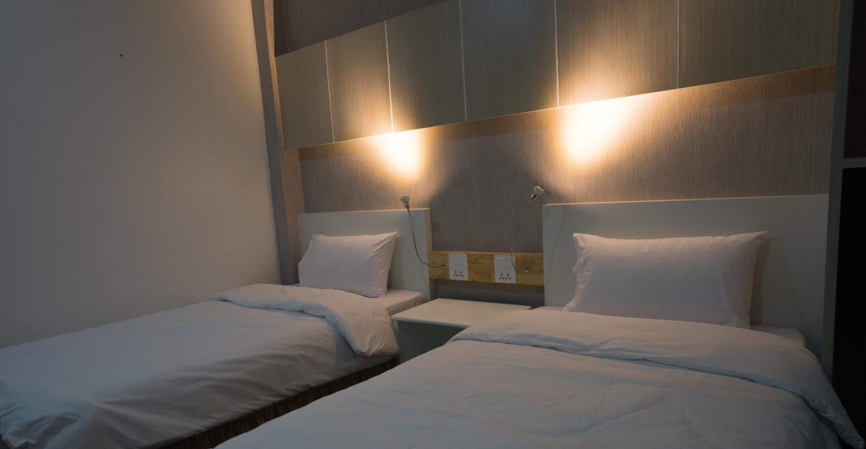 Habitación de hotel con dos camas individuales, cabecero de madera y lámpara fijada a la pared.