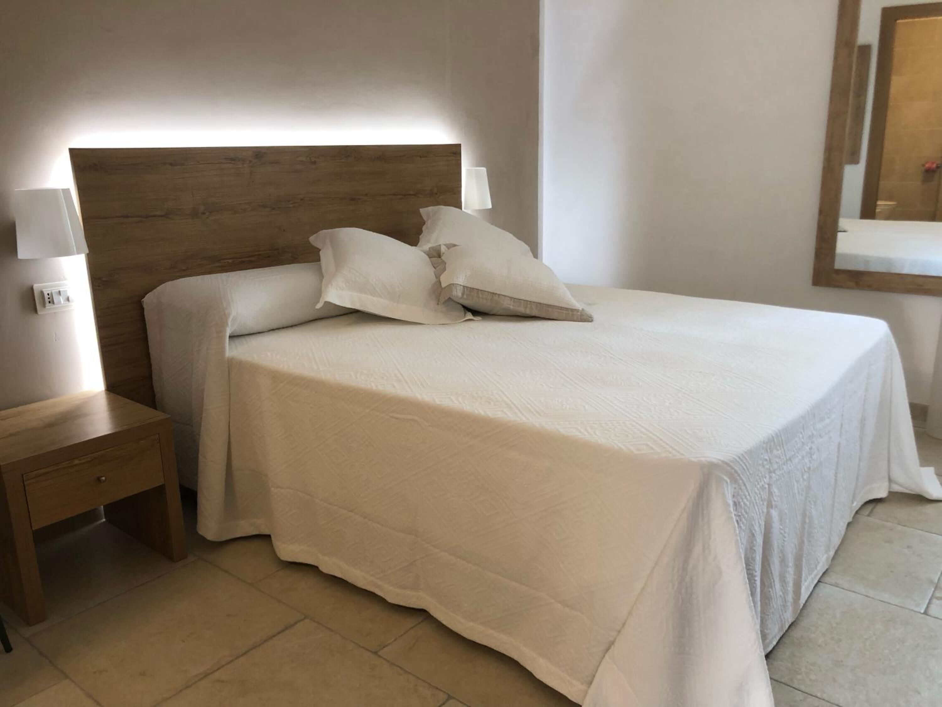 Ein ordentlich bezogenes Bett mit Holzkopfschild, ein Nachttisch mit Lampe und ein Spiegel an der Wand.