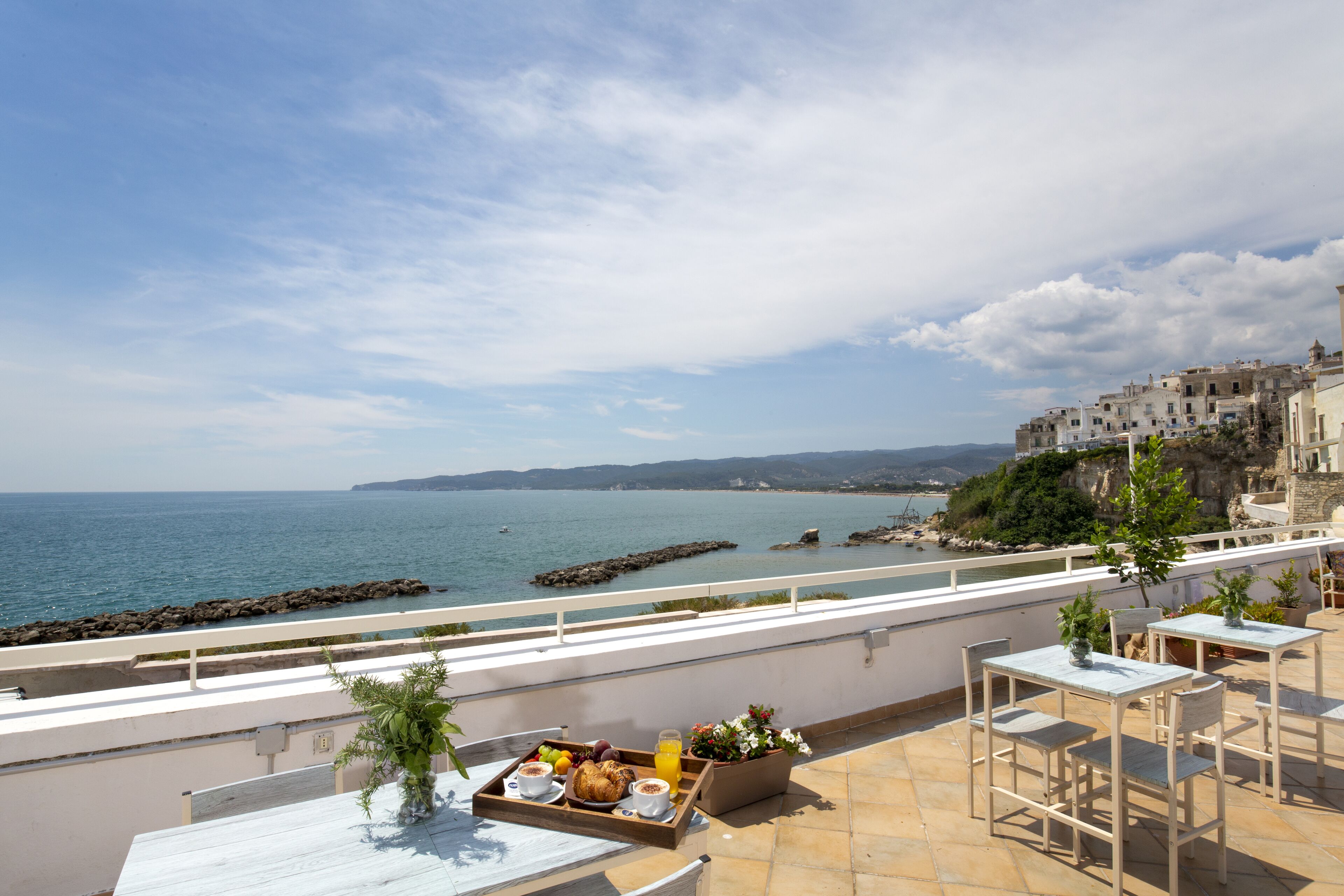 Une terrasse avec une table dressée pour le petit-déjeuner, donnant sur la mer, une balustrade blanche et une vue sur les bâtiments qui bordent la côte.