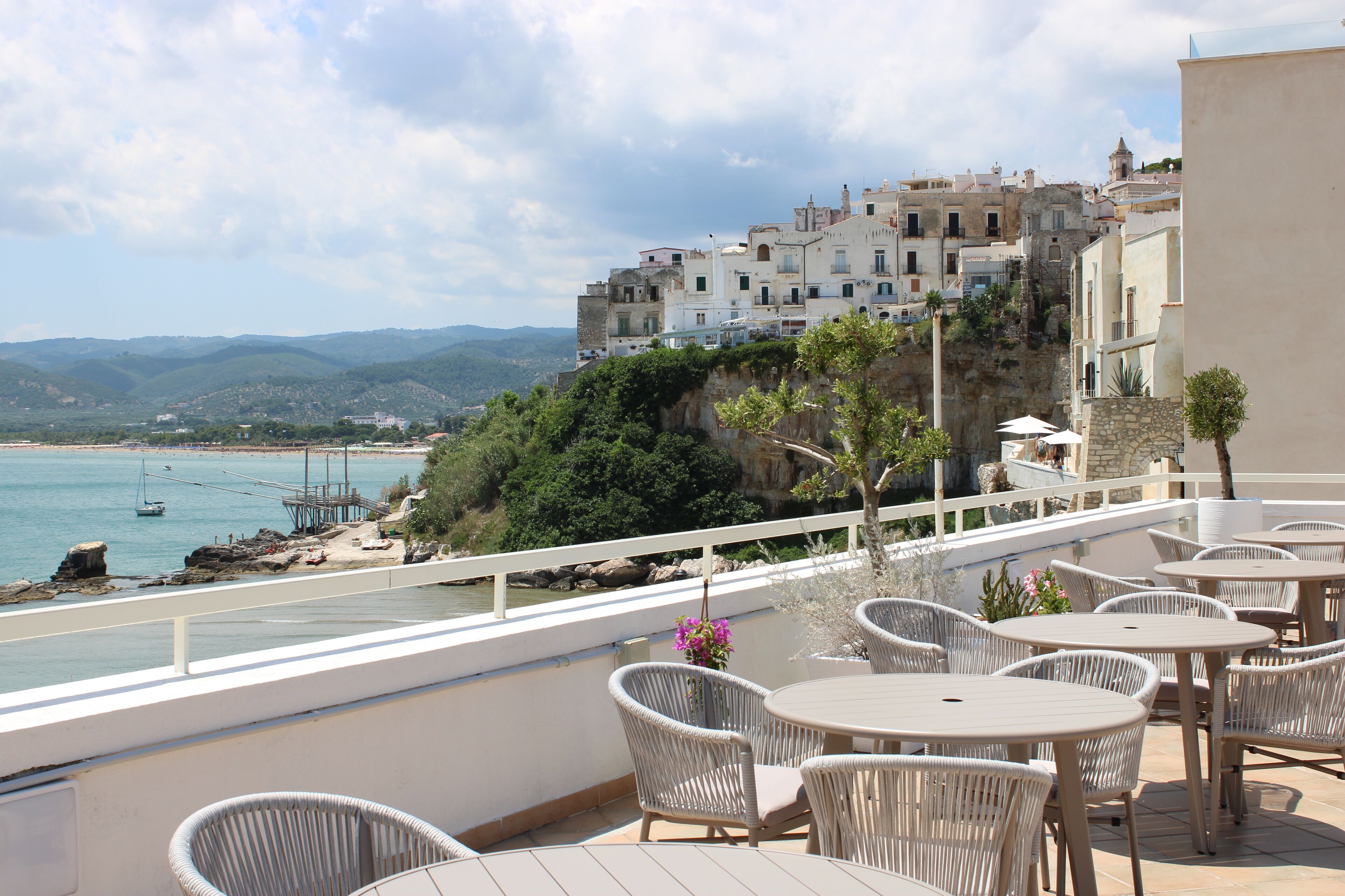 A terrace with wicker chairs and tables overlooking a coastal town with buildings on a hillside.