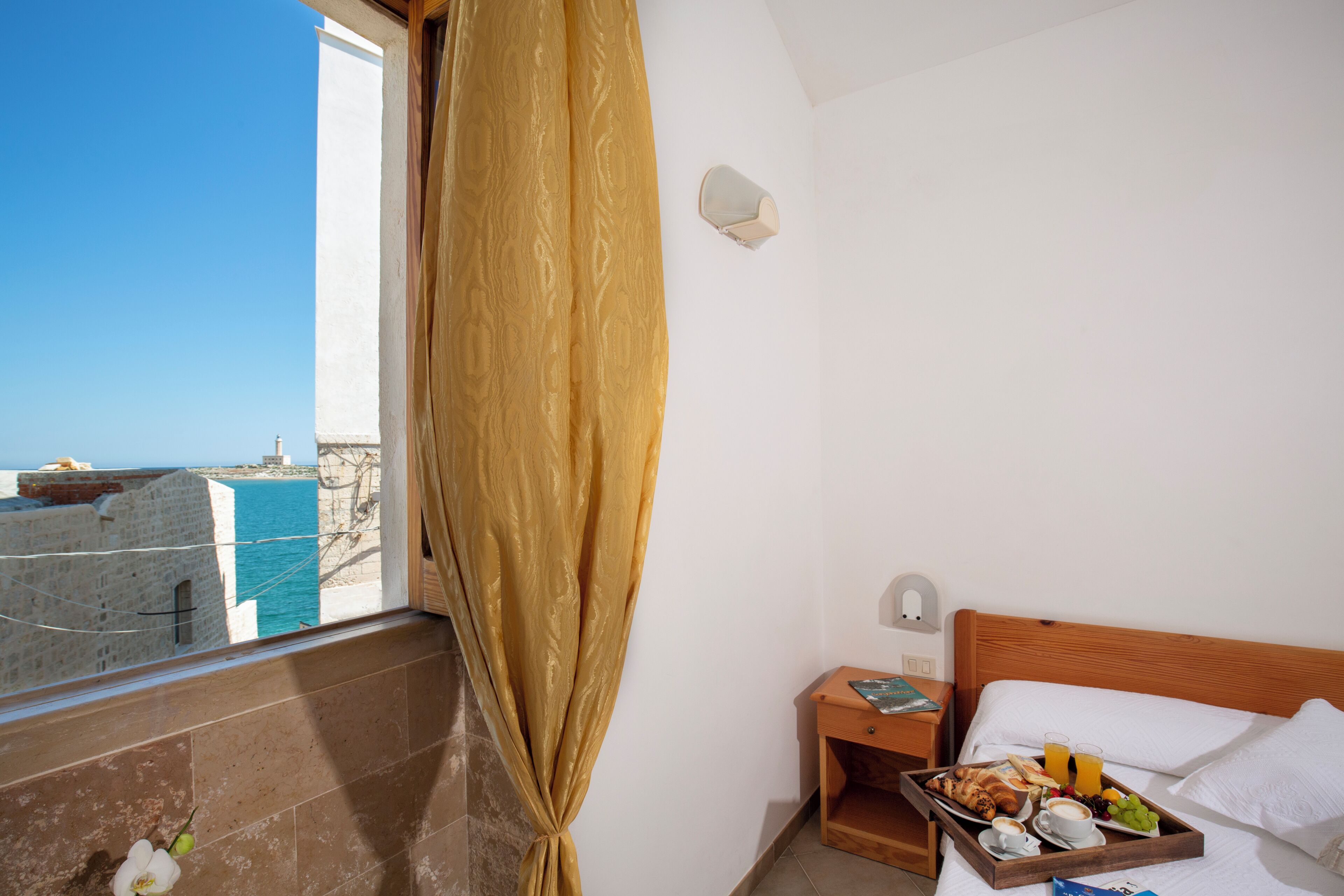 A hotel room with a bed, a wooden nightstand, a tray with breakfast, a window with a curtain, and a view of the sea.