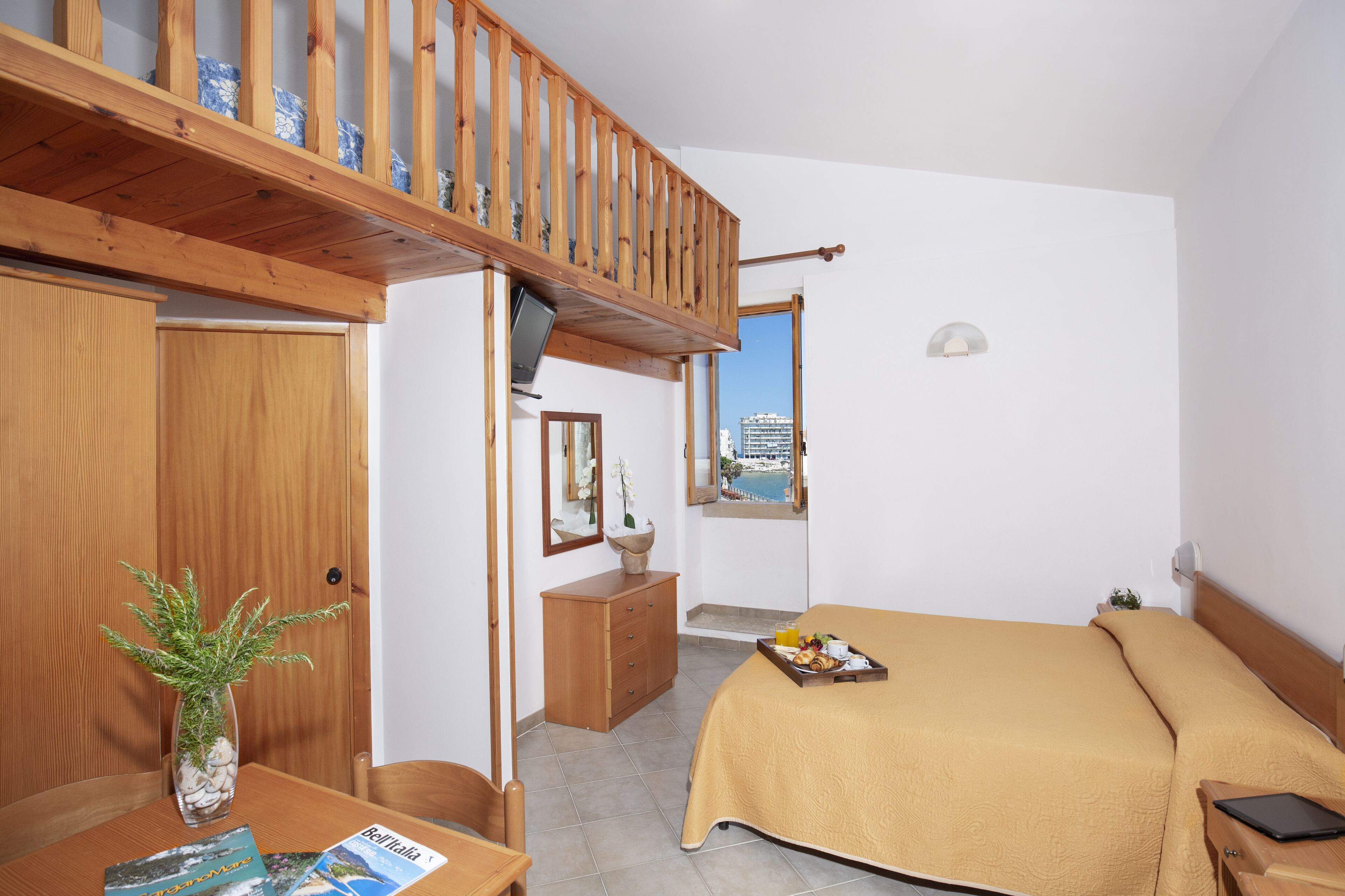 Une chambre à coucher comprenant un lit, un bureau en bois, un vase avec des plantes, une fenêtre donnant sur la mer et un balcon avec des balustrades en bois.