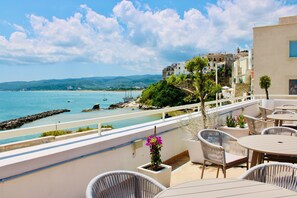 Eine Terrasse mit Korbmöbeln, Meerblick und Gebäuden im Hintergrund.