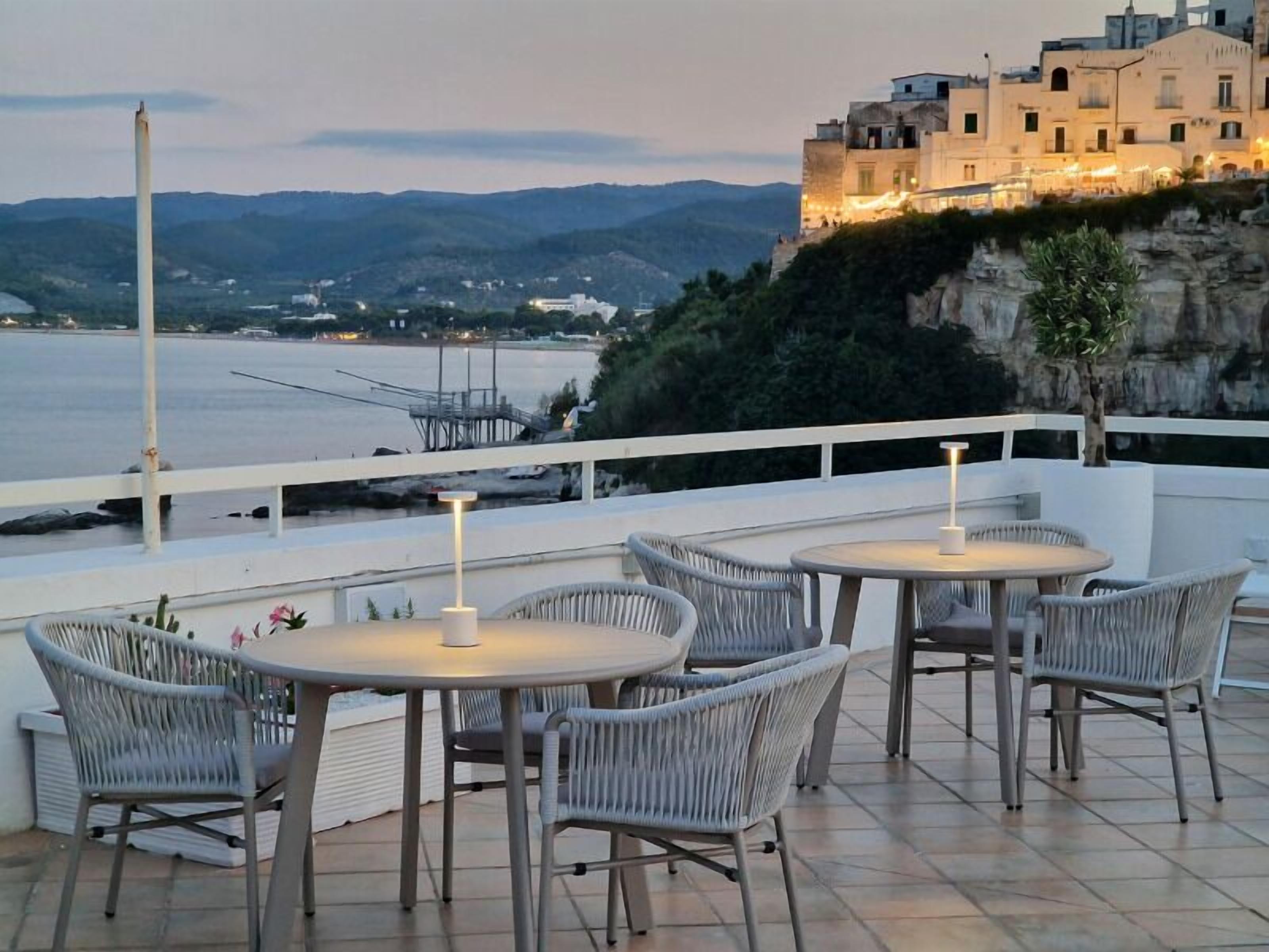 A terrace with wicker chairs and tables, overlooking a body of water and a mountainous landscape.