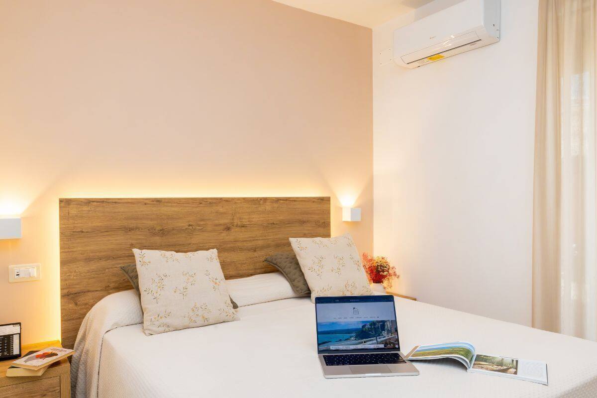 Una cama bien hecha con un ordenador portátil y una revista, una mesita de noche con un teléfono y un libro, y un equipo de aire acondicionado en la pared.