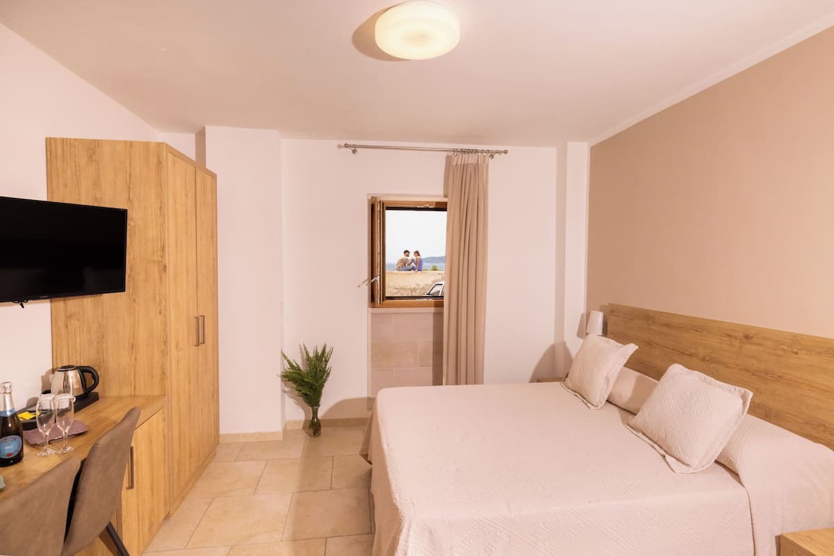 Habitación de hotel con cama, escritorio con televisor de pantalla plana, pequeña mesa con planta y vista a la playa a través de una puerta abierta.