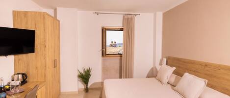 A hotel room with a bed, a desk with a flat-screen TV, a small table with a plant, and a view of the beach through an open door.