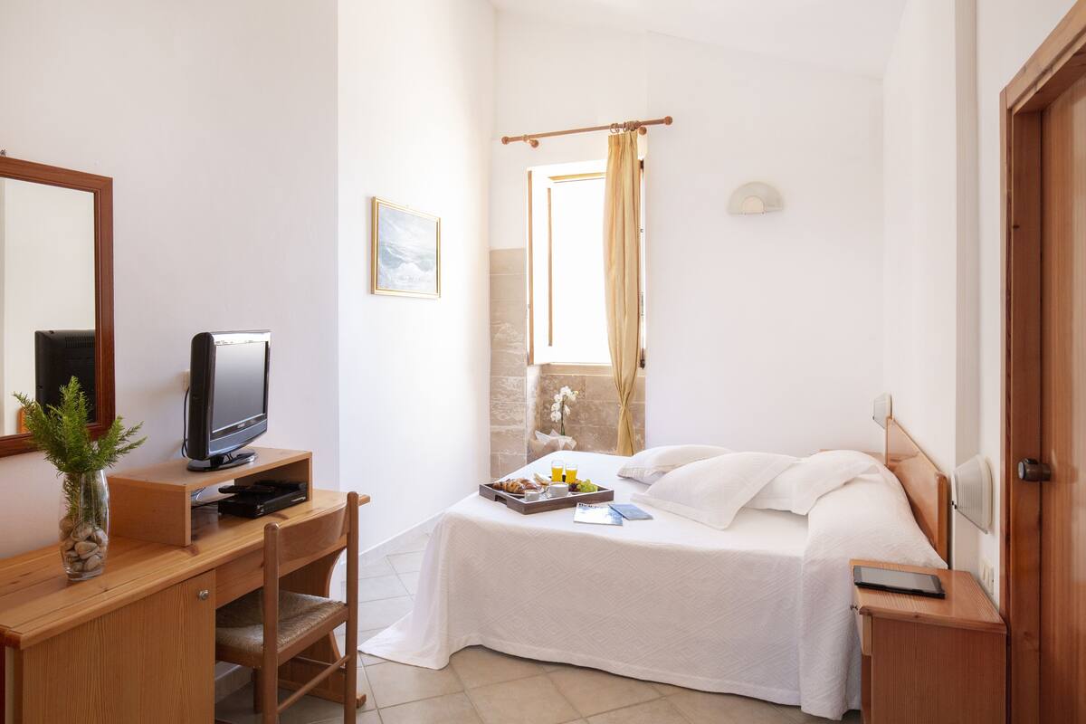 Habitación de hotel con cama, escritorio con televisor, pequeña mesa con desayuno, ventana con cortinas y espejo.